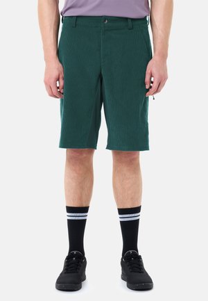 Uomo che indossa pantaloncini teal fino al ginocchio, calze nere a coste con strisce bianche e scarpe da ginnastica nere, in piedi su uno sfondo bianco.