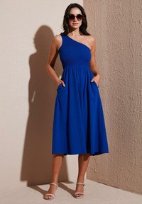 Blue one-shoulder dress with a smocked bodice, flowing skirt, and pockets. Smooth texture and mid-calf length, paired with white heels.