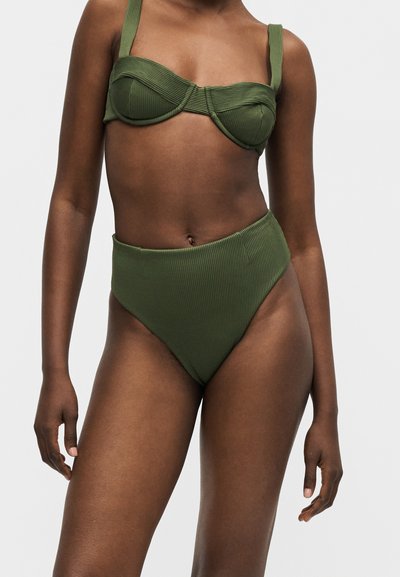 Femme portant un bikini côtelé vert olive avec un haut à armatures et un bas taille haute sur fond gris uni.