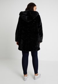Black faux fur coat with a hood, featuring a loose fit and long sleeves, paired with dark jeans and white sneakers. Smooth texture throughout.