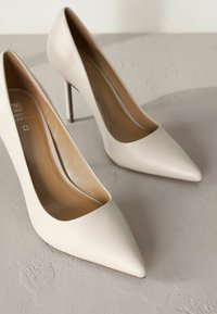Beige korkokenkiä, joissa on sileä nahkapinta, terävä kärki ja tyylikäs stiletto-korko, asetettuna tekstuurimaiselle harmaalle pinnalle.