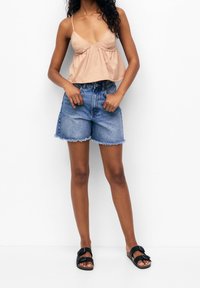 Top senza maniche di un beige chiaro con un design svasato, abbinato a pantaloni shorts in denim blu a vita alta con orlo sfrangiato. Indossato con sandali neri.