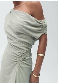 Schulterfreies, hellgrünes Kleid mit gerafften Ärmeln und gerafften Seiten; umfasst ein goldenes Armband. Weicher Stoff mit glatter Textur.