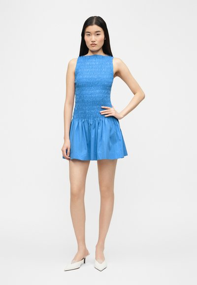 Νεαρή γυναίκα με μακριά μαύρα μαλλιά, φορώντας ένα αμάνικο μπλε μίνι φόρεμα με υφή και λευκά μυτερά τακούνια, μπροστά σε απλό φόντο.