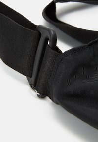 Ceinture en tissu noir avec une surface texturée, équipée d'une boucle en plastique et d'un anneau en métal. Le design est utilitaire et réglable.