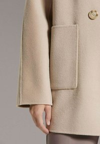 Beige wollen jas met een gestructureerde vorm, voorzien van een grote opgestikte zak en een ronde knoopsluiting aan de bovenkant van de mouw.