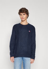 Pull en tricot bleu marine avec un col rond, des poignets et un ourlet côtelés. Présente un petit logo multicolore sur la poitrine. Design texturé.