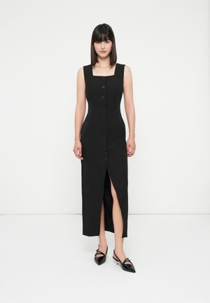 Femeie stând în picioare, purtând o rochie midi neagră, cu nasturi, fără mâneci, care are o fendere în față, și tocuri slingback negre, pe un fundal alb.