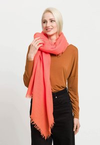 Korallenfarbener Schal, der um den Hals gewickelt ist, aus weichem Stoff mit fransigen Kanten, kombiniert mit einem braunen Langarmoberteil und schwarzen Hosen.