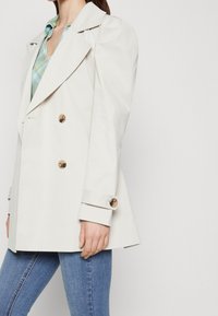 Abrigo trench de doble botonadura en color crema, con grandes botones, un amplio cuello y mangas ajustadas, llevado sobre una camisa de cuadros verde y unos vaqueros.