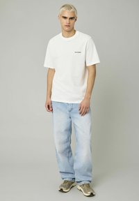 T-shirt en coton blanc avec le texte "Untroubled", associé à un jean bleu clair et large, et des baskets grises, sur un fond neutre.