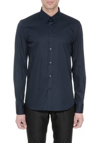 Homme portant une chemise cintrée bleu foncé à manches longues boutonnée et un pantalon noir, debout devant un fond blanc.