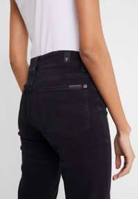 Jeans negros con cintura alta, corte ajustado y bolsillos estándar. Detalles de costura destacados en los bolsillos traseros y una pequeña etiqueta de la marca.