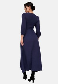 Langes, marineblaues Kleid mit gerafftem Oberteil und Puffärmeln. Fließender Stoff und Saum bis zum Knöchel. Kombiniert mit schwarzen Stiefeln mit Absatz.