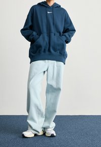 Persona vistiendo una sudadera de Nike de color azul marino, jeans sueltos de color azul claro y zapatillas blancas, de pie sobre una alfombra azul contra una pared blanca lisa.