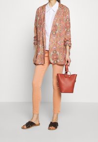 Blazer en tissu léger à motif floral rose et crème, chemise blanche à col, jean skinny pêche, sandales marron à enfiler, sac seau rouge.