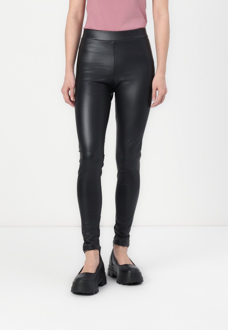 Personne debout portant des leggings noirs en simili cuir, un haut rose et des chaussures à plateforme noires épaisses sur un fond clair uni.