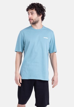 Homme aux cheveux bouclés portant un t-shirt bleu clair ATHENA et un short noir, regardant vers sa gauche sur un fond uni.