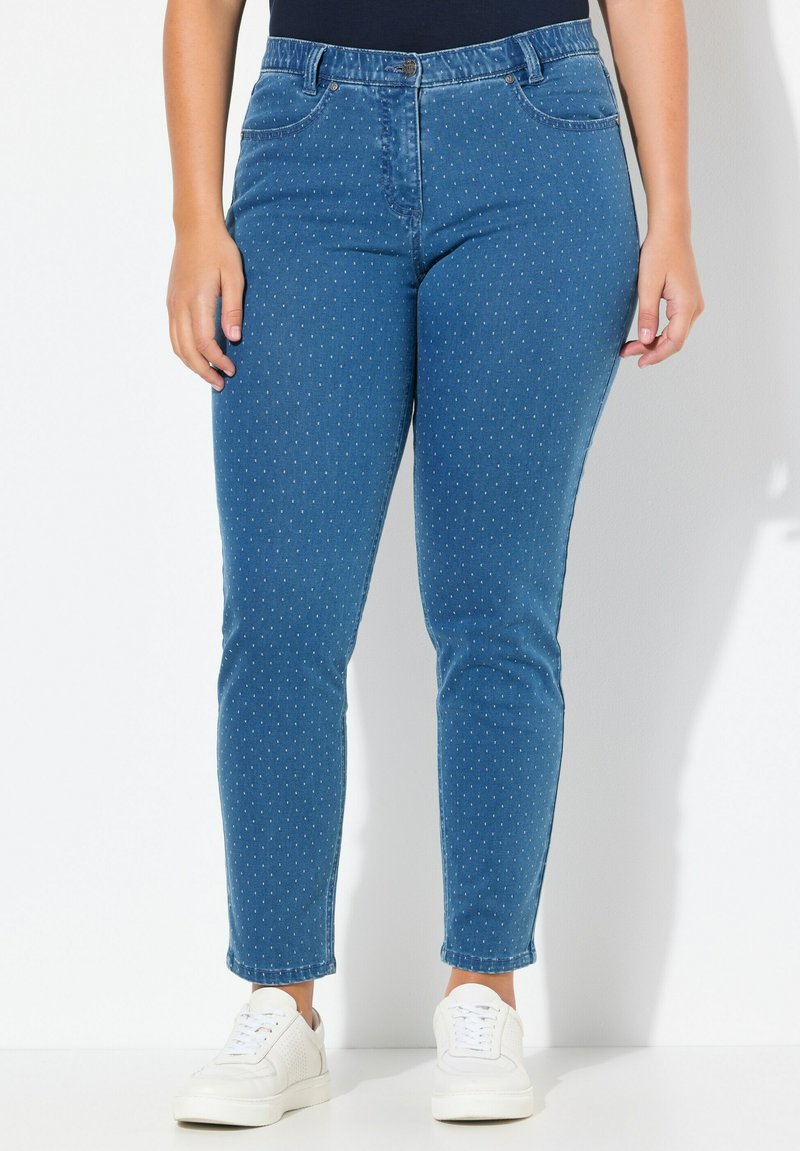 Vaqueros de mezclilla azul con pequeños lunares blancos, corte ajustado y diseño de cinco bolsillos, combinados con zapatillas blancas. Textura suave.