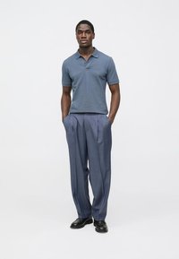 Tumši zils polo krekls kombinācijā ar gaiši ziliem krokotiem biksēm. Apģērbam ir ikdienišķs piegriezums un gludas tekstūras, ko papildina melnas kurpes.