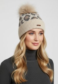 Blonde woman with wavy hair wearing a beige leopard print knit hat with a pom-pom and a dark gray ribbed turtleneck sweater.