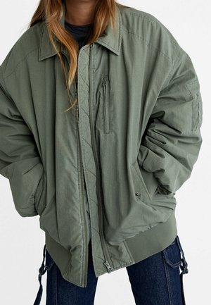 Vrouw draagt een oversized olijfgroene bomberjas met ritssluiting en zakken, gecombineerd met donkerblauwe spijkerbroek.