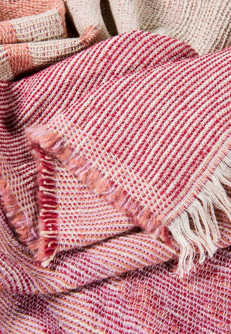 Gewebte Decke in den Farbtönen Rosa und Rot, mit einem strukturierten Muster und fransigen Kanten. Weiches Material mit einem gemütlichen Erscheinungsbild.