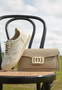 Béžové tenisky s texturovanými panely a světlými tkaničkami, vedle béžové kožené tašky s výrazným zapínáním, položené na židli.