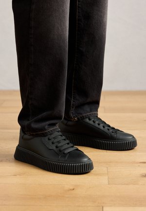 Person wearing black ribbed sole sneakers and black jeans standing on light wooden floor.