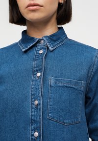 Femme portant une chemise en denim bleue à boutons-pression argentés et poche poitrine, montrée des épaules aux lèvres.