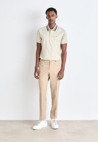Beige polo shirt met zwart-wit gestreepte kraag, gecombineerd met lichtbruine op maat gemaakte broek en witte sneakers. Eenvoudig ontwerp, slim fit.