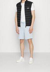 Svart pufferväst med horisontell sömnad, vit dragkedja och "BOSS"-logotyp, bärs över en vit t-shirt, i kombination med ljusblå shorts och vita sneakers.