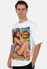 Wit katoenen T-shirt met een kleurrijke afbeelding van een zittende vrouw in zwemkleding met de tekst "Good Vibes"; levendige achtergrond met strepen en spatten.