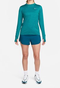 Teal long-sleeve athletic shirt with thumbholes, paired with teal shorts. Worn with white and teal sneakers, featuring a reflective logo.