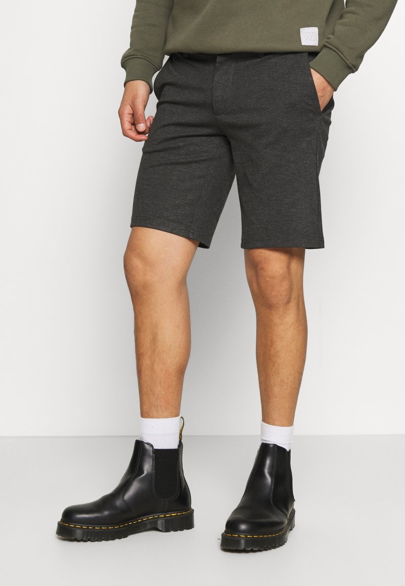 Pantaloni corti grigio carbone realizzati in materiale morbido con fronte piatto, abbinati a stivali neri alla caviglia con cuciture gialle e pannelli elasticizzati laterali.