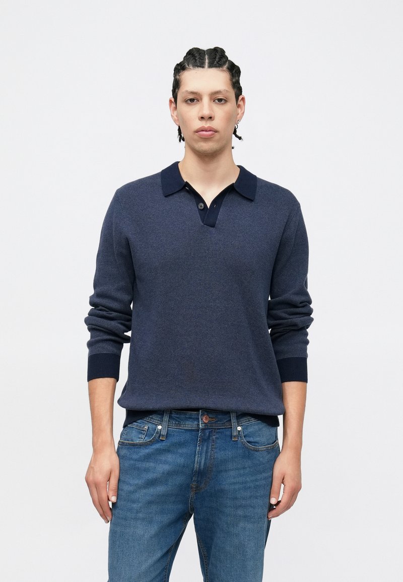 Marineblauer Strick-Polo-Pullover mit kontrastierendem Kragen, zwei Knöpfen und gerippten Bündchen. Kombiniert mit hellblauen Jeans.