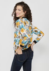 Giacca bomber floreale con base bianca, caratterizzata da fiori gialli e foglie turchesi; colletto e polsini a coste neri; tessuto leggero.