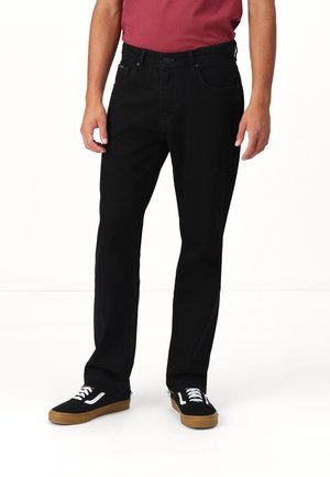 Mann trägt schwarze Jeans mit geradem Bein, schwarze und weiße Sneaker mit Kautschuksohlen und ein rotes Hemd, steht vor weißem Hintergrund.