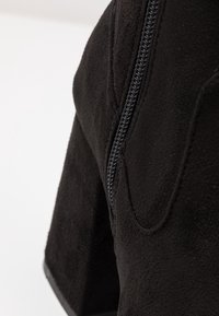 Primer plano de material de ante negro, con una cremallera plateada prominente a lo largo del costado y una superficie texturizada con un acabado suave.