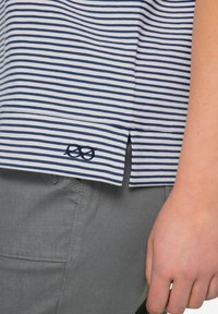 Pasiasta koszula z krótkim rękawem w kolorze białym i granatowym, z bocznym rozcięciem i haftowanym detalem w kształcie pętli blisko dołu; zestawiona z jasnoszarymi spodniami.