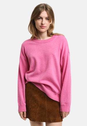 Růžový oversized svetr z měkké tkaniny s kulatým výstřihem, dlouhými rukávy a uvolněným střihem, v kombinaci s hnědou sukní z semiše.