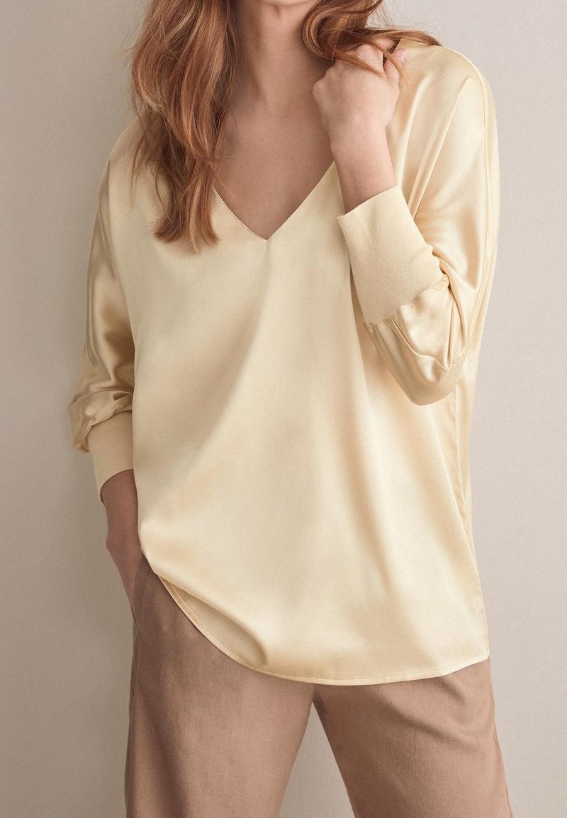 Blouse satin couleur crème avec un décolleté en V et des manches longues. Texture lisse, coupe décontractée et détails de manches à poignets. Associée à un pantalon beige.