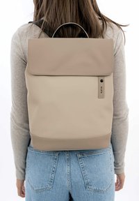 Zaino beige realizzato in materiale sintetico liscio, con un cappuccio superiore, un'etichetta con logo discreta e un fondo arrotondato. Design semplice con una palette di colori monocromatici.