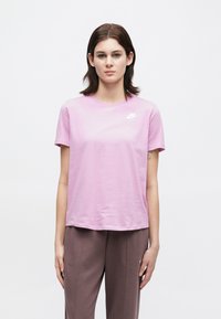 Γυναίκα που στέκεται μπροστά σε λευκό φόντο, φορώντας ροζ μπλούζα Nike και καφέ παντελόνι, κοιτάζοντας μπροστά με τα χέρια χαλαρά στα πλάγια.