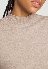 Beiger Strickpullover mit hohem, geripptem Kragen und weicher Textur, der eine Nahaufnahme des subtilen Musters des Stoffes zeigt.