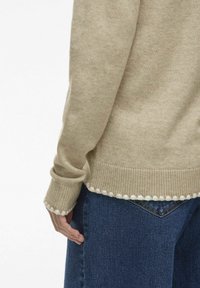 Suéter de punto beige con puños acanalados y un borde con puntas en el dobladillo, combinado con denim azul oscuro. Textura suave, diseño casual.
