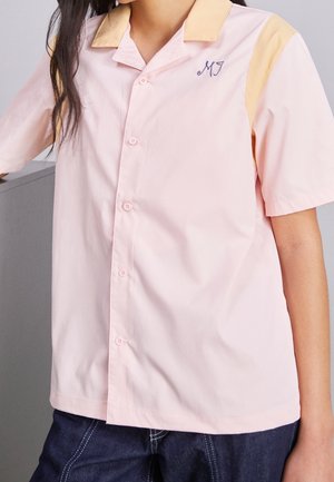 Personne portant une chemise à manches courtes, boutonnée, de couleur rose clair avec des accents jaunes sur le col et les épaules, avec les initiales brodées "MJ" sur la poitrine.