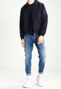 Námořnická bomber bunda se zipem vpředu, žebrovanými manžetami a dvěma bočními kapsami. Světle modré džíny, ohrnuté na kotnících, a bílé tenisky.