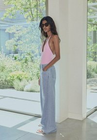 Pink deep-V swimsuit, light blue wide-leg jeans, and sunglasses. Model stands in a bright room with greenery visible through large windows.