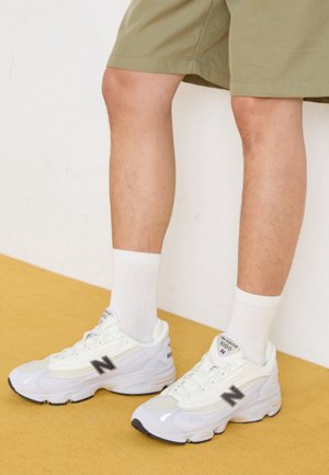 Jambes portant un short beige, des chaussettes blanches montantes et des baskets blanches New Balance 1000, debout sur un sol jaune contre un mur blanc.